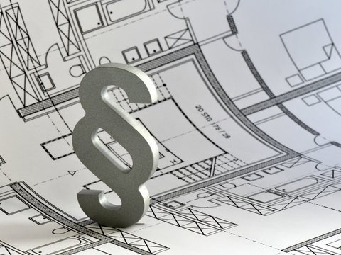 Ein silbernes Paragrafenzeichen steht auf einem ausgebreiteten architektonischen Grundrissplan