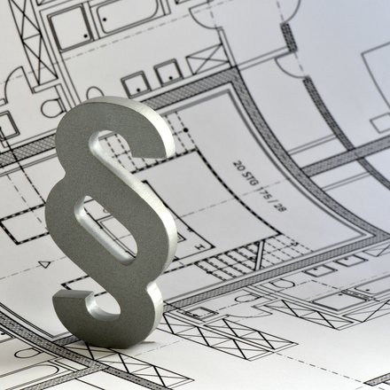 Ein silbernes Paragrafenzeichen steht auf einem ausgebreiteten architektonischen Grundrissplan