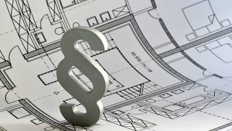 Ein silbernes Paragrafenzeichen steht auf einem ausgebreiteten architektonischen Grundrissplan
