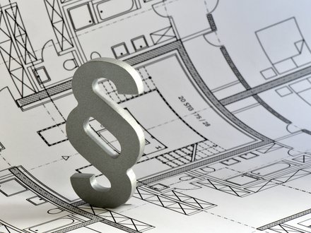 Ein silbernes Paragrafenzeichen steht auf einem ausgebreiteten architektonischen Grundrissplan