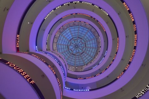 Blick nach oben durch ein spiralförmiges Treppenhaus mit einer Glaskuppel und leuchtenden Textbändern an den Geländern