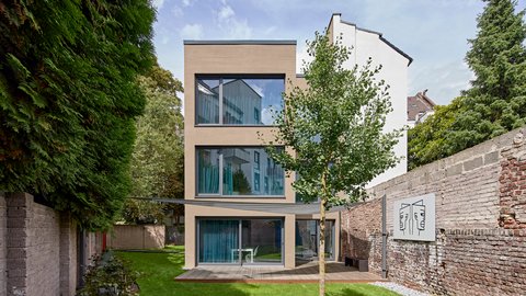 Modernes dreigeschossiges Wohnhaus mit heller Fassade, großen bodentiefen Fenstern und Holzterrasse, umgeben von Garten, Bäumen und historischen Backsteinmauern