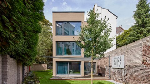 Modernes dreigeschossiges Wohnhaus mit heller Fassade, großen bodentiefen Fenstern und Holzterrasse, umgeben von Garten, Bäumen und historischen Backsteinmauern
