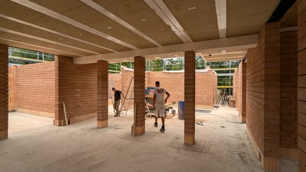Innenaufnahme Rohbau: Rote Ziegelwände und -stützen, Decke mit Holzträgern; zwei Bauarbeiter zwischen Werkzeugen und Leiter, Tageslicht fällt durch offene Bereiche.