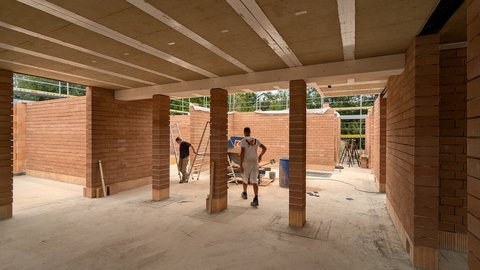 Innenaufnahme Rohbau: Rote Ziegelwände und -stützen, Decke mit Holzträgern; zwei Bauarbeiter zwischen Werkzeugen und Leiter, Tageslicht fällt durch offene Bereiche.