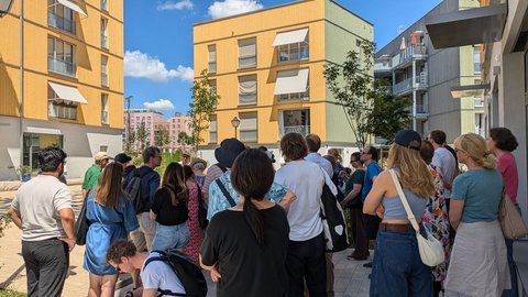 Gruppe von Menschen bei einer Architekturführung in einem modernen Wohnquartier mit gelben Holzfassaden.