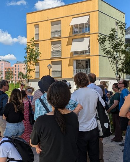 Gruppe von Menschen bei einer Architekturführung in einem modernen Wohnquartier mit gelben Holzfassaden.