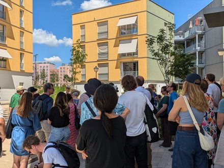 Gruppe von Menschen bei einer Architekturführung in einem modernen Wohnquartier mit gelben Holzfassaden.