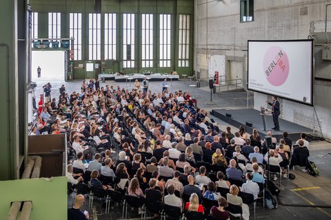 Publikum verfolgt einen Vortrag in einer großen Industriehalle mit Präsentationsleinwand.