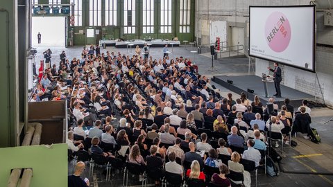 Publikum verfolgt einen Vortrag in einer großen Industriehalle mit Präsentationsleinwand.