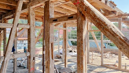 Offengelegte Fachwerkkonstruktion auf sonniger Baustelle, grob behauene Balken mit Markierungen; Dorfhäuser hinten.