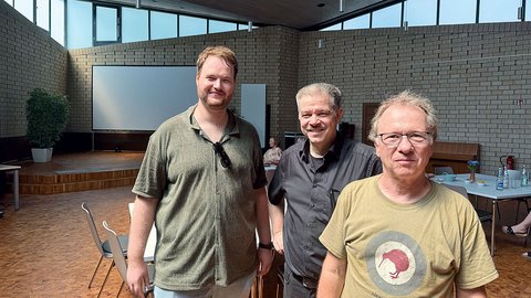 Drei Männer stehen in einem hellen Raum mit großen Fenstern und Stühlen, im Hintergrund eine Leinwand und eine Pflanze