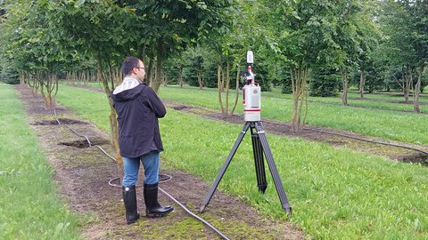 Mann in schwarzer Jacke und Gummistiefeln bedient auf Waldweg ein Laserscanner-Messgerät auf Stativ. Das Gerät ist weiß und rot gefärbt und auf einen Baum ausgerichtet. Hinter ihm verlaufen Reihen junger Bäume mit Bewässerungsleitungen auf feuchtem Waldboden. Grüne Wiese säumt die gepflasterten Wege.