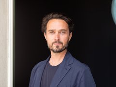 Portrait eines Mannes mit dunklem, leicht gewelltem Haar, Dreitagebart und blauem Sakko vor schwarzem Hintergrund.