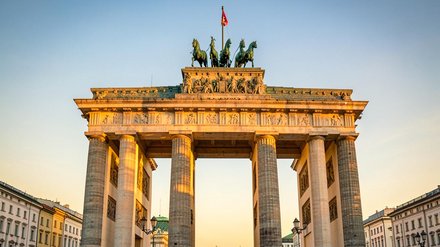 Frontale Aufnahme des Brandenburger Tors in Berlin bei warmem Abendlicht, Menschen und Fahnen im Vordergrund, goldene Quadriga auf dem Scheitel sichtbar.