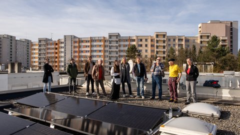 "Eine Gruppe von etwa zehn Personen steht nebeneinander auf einem Flachdach. Im Vordergrund liegen flache Solarmodule. Im Hintergrund reihen sich bei strahlendem Sonnenschein mehrstöckige Wohnblöcke aneinander."