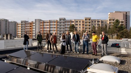 "Eine Gruppe von etwa zehn Personen steht nebeneinander auf einem Flachdach. Im Vordergrund liegen flache Solarmodule. Im Hintergrund reihen sich bei strahlendem Sonnenschein mehrstöckige Wohnblöcke aneinander."