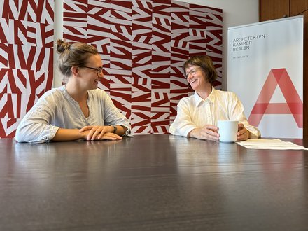  Zwei Frauen sitzen einander an einem dunklen Tisch gegenüber und unterhalten sich lachend. Die Frau links trägt eine gestreifte Bluse, die Frau rechts eine weiße Bluse und hält eine weiße Tasse. Im Hintergrund ist eine Wand mit einem rot-weißen geometrischen Muster sowie ein Roll-up-Banner der Architektenkammer Berlin zu sehen.