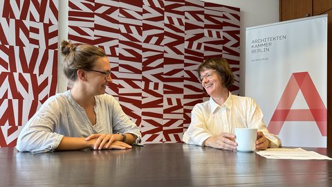  Zwei Frauen sitzen einander an einem dunklen Tisch gegenüber und unterhalten sich lachend. Die Frau links trägt eine gestreifte Bluse, die Frau rechts eine weiße Bluse und hält eine weiße Tasse. Im Hintergrund ist eine Wand mit einem rot-weißen geometrischen Muster sowie ein Roll-up-Banner der Architektenkammer Berlin zu sehen.