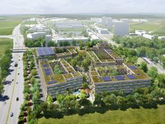 "Luftaufnahme einer modernen Wohnsiedlung in ländlicher Umgebung. Im Vordergrund mehrere vier- bis fünfgeschossige Wohngebäude mit begrünten Dächern, Solarpanelen und integrierten Grünflächen. Die Gebäude sind in Holz- und hellen Steinoptik gestaltet. Ein Spielplatz mit rotem Spielgerät ist zwischen den Gebäuden sichtbar. Dahinter erstrecken sich grüne Wiesen und Bäume. Im Hintergrund große Industriehallen und Gewerbegebäude mit weißen Fassaden. Links eine Hauptstraße mit Fahrzeugen und Straßenbäumen. Die Umgebung ist geprägt von grünen Feldern und vereinzelten Baumgruppen unter bewölktem Himmel."