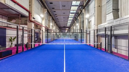 Lange, hohe Industriehalle mit mehreren Padelplätzen in Reihe; leuchtend blaue Spielflächen mit Netzen, Glas- und Gitterwänden. Seitlich pinke Laufwege, oben Oberlichter und rote Leitungen; keine Personen.