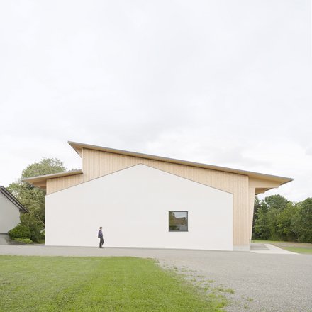 Moderne Gemeindehalle mit weißer Fassade und Holzverkleidung, davor eine Person auf einem Gehweg, umgeben von Rasen und Bäumen