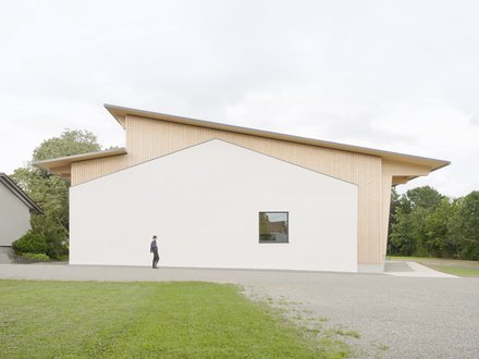 Moderne Gemeindehalle mit weißer Fassade und Holzverkleidung, davor eine Person auf einem Gehweg, umgeben von Rasen und Bäumen