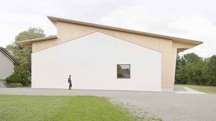 Moderne Gemeindehalle mit weißer Fassade und Holzverkleidung, davor eine Person auf einem Gehweg, umgeben von Rasen und Bäumen