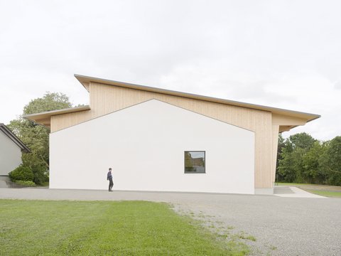Moderne Gemeindehalle mit weißer Fassade und Holzverkleidung, davor eine Person auf einem Gehweg, umgeben von Rasen und Bäumen