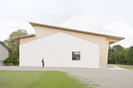 Moderne Gemeindehalle mit weißer Fassade und Holzverkleidung, davor eine Person auf einem Gehweg, umgeben von Rasen und Bäumen