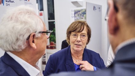 Nahaufnahme in heller Ausstellung. Die Frau blickt sprechend zur Kamera, Hand am Revers. Zwei Gesprächspartner sind von hinten angeschnitten. Hinter ihr stehen Infotafeln mit Logos „Baukultur.SH“ und „Schleswig‑Holstein“.