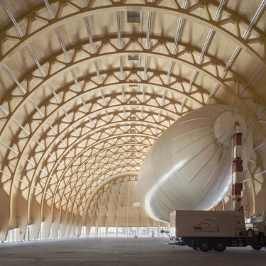 Großer Holzbau-Hangar mit geschwungenem Tragwerk und einem Luftschiff im Innenraum.