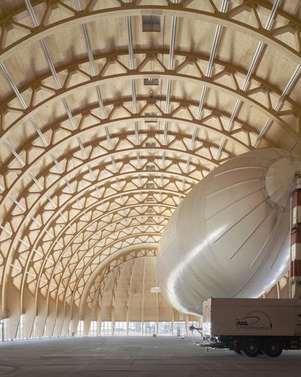 Großer Holzbau-Hangar mit geschwungenem Tragwerk und einem Luftschiff im Innenraum.