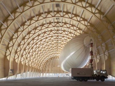 Großer Holzbau-Hangar mit geschwungenem Tragwerk und einem Luftschiff im Innenraum.