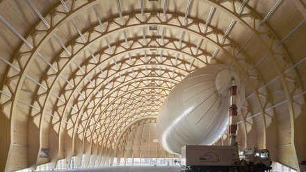 Großer Holzbau-Hangar mit geschwungenem Tragwerk und einem Luftschiff im Innenraum.