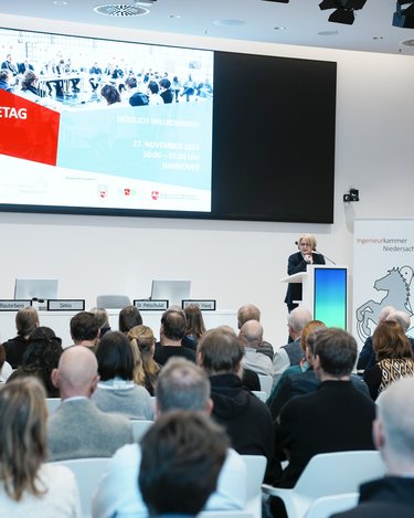 Blick in einen voll besetzten Veranstaltungssaal mit Podium, Rednerpult und großer Präsentationsleinwand.