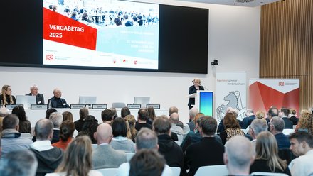 Blick in einen voll besetzten Veranstaltungssaal mit Podium, Rednerpult und großer Präsentationsleinwand.