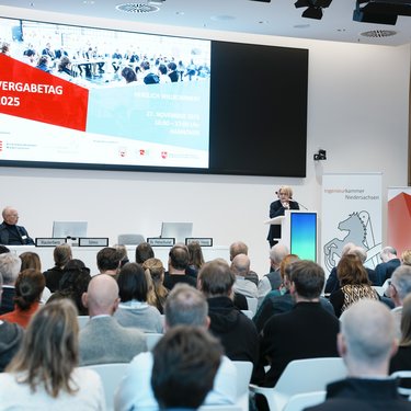 Blick in einen voll besetzten Veranstaltungssaal mit Podium, Rednerpult und großer Präsentationsleinwand.