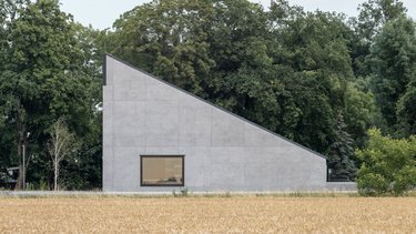 Minimalistisches Betongebäude mit schrägem Dach am Rand eines Getreidefeldes.