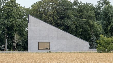 Minimalistisches Betongebäude mit schrägem Dach am Rand eines Getreidefeldes.