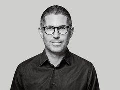 Schwarzweiß‑Porträt einer Person mit runder Brille vor grauem Hintergrund.