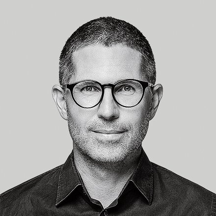 Schwarzweiß‑Porträt einer Person mit runder Brille vor grauem Hintergrund.