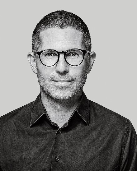 Schwarzweiß‑Porträt einer Person mit runder Brille vor grauem Hintergrund.