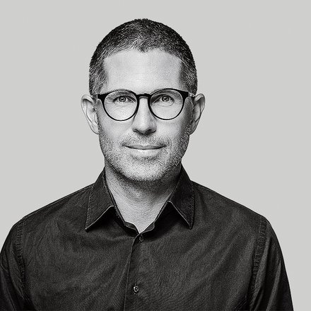 Schwarzweiß‑Porträt einer Person mit runder Brille vor grauem Hintergrund.