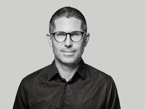 Schwarzweiß‑Porträt einer Person mit runder Brille vor grauem Hintergrund.
