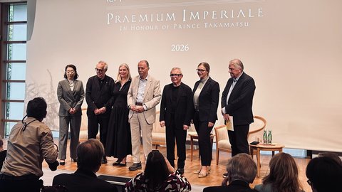 Sieben Personen stehen nebeneinander auf einer Bühne vor einer Leinwand mit der Aufschrift 'PRAEMIUM IMPERIALE IN HONOUR OF PRINCE TAKAMATSU 2026', Publikum im Vordergrund.