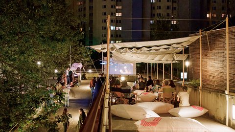 "Nächtliche Szene bei einem Freiluftkino. Unter einem hellen Stoffdach sitzen Menschen auf großen weißen Sitzsäcken und Liegestühlen und schauen auf eine beleuchtete Leinwand. Im dunklen Hintergrund ragen Hochhäuser auf."