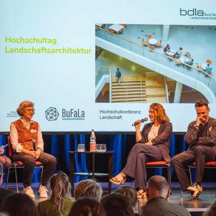 Podiumsdiskussion auf einer Bühne: fünf Personen sitzen auf roten Stühlen, eine spricht ins Mikrofon. Dahinter Leinwand mit „Hochschultag Landschaftsarchitektur“, Logos von Verbänden und Foto einer Treppenhalle.