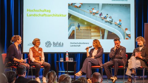 Podiumsdiskussion auf einer Bühne: fünf Personen sitzen auf roten Stühlen, eine spricht ins Mikrofon. Dahinter Leinwand mit „Hochschultag Landschaftsarchitektur“, Logos von Verbänden und Foto einer Treppenhalle.