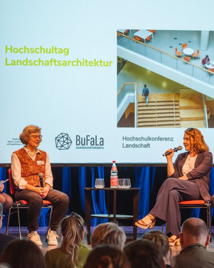 Podiumsdiskussion auf einer Bühne: fünf Personen sitzen auf roten Stühlen, eine spricht ins Mikrofon. Dahinter Leinwand mit „Hochschultag Landschaftsarchitektur“, Logos von Verbänden und Foto einer Treppenhalle.